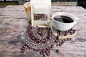 Full of coffee beans spilling out bag on brown wooden background with a cup of black coffee