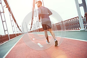 Full of energy. Back view of young handsome man in sports clothing jogging on the bridge in the morning