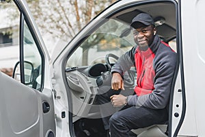 Full of energy, positive African-American deliveryman posing on driver's seat with open door. Courier outfit - black