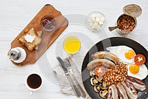 Full English breakfast in a pan with fried eggs, bacon, beans, sausages and toasts on white wooden background, top view. From abo