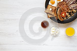 Full English breakfast in a pan with fried eggs, bacon, sausages, beans, toasts and orange juice on white wooden background with c