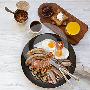 Full English breakfast in a pan with fried eggs, bacon, sausages, beans and toasts on white wooden table, top view. Flat lay. From