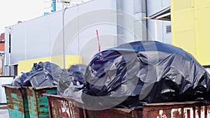 Full garbage containers with large black bags of garbage in wind close-up, near an industrial building. Environmental