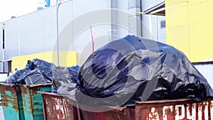 Full garbage containers with large black bags of garbage in wind close-up, near an industrial building. Environmental