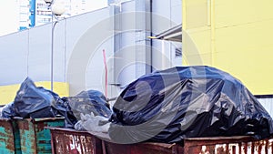 Full garbage containers with large black bags of garbage in wind close-up, near an industrial building. Environmental