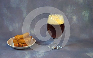 A full glass with dark beer and foam and wheat croutons on a plate