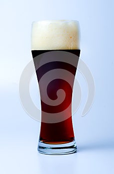 A full glass of dark beer with a large cap of froth. Slightly bluish background. Side view