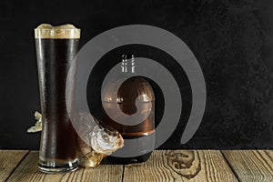Full glass of dark foamy beer with dried fish and a bottle on rough boards on a black concrete background. Artistic mockup with