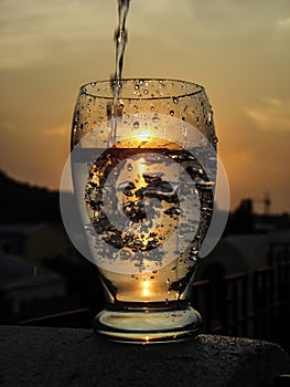 A full glass of water with a pouring waterjet and a sun downing