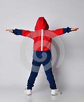 Full growth portrait of frolic blond kid boy in red and blue sport suit standing back to us with arms outstretched