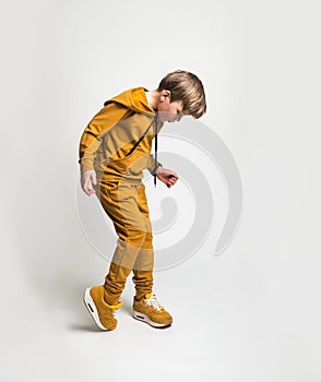 Full growth portrait of stylish cool blond kid boy in yellow sport suit hoodie and pants. Frolic child with hands in