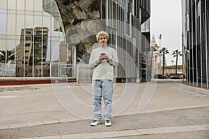 In full growth young caucasian guy with curly blonde hair using smartphone at city centre.