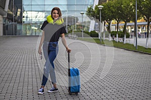 Full height portrait of young beautiful woman in blue jeans and black t-shirt with blue suitcase and yellow travel pillow.