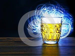 Full jar of lager beer with froth on a wooden table glowing in a dark, blue light painting in the background behind the glass.