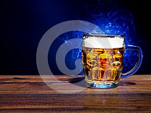 Full jar of lager beer with froth on a wooden table glowing in a dark, blue light painting in the background behind the glass.