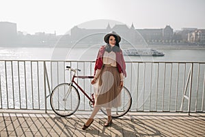 Full-legth portrait of gorgeous woman in vintage clothes posing with pleasure near river. Outdoor shot of appy brunette