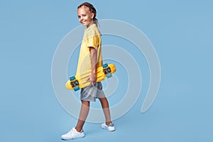 Full lengh image of stylish little boy with african dreads with headphone and skateboard posing over blue background.