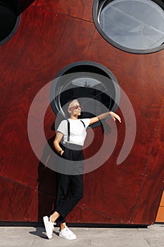 Full lengh photo of hipster young woman wearing red sunglasses and black hat posing near wall of modern building.