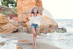 Full-lengh photo of pretty blonde girl with long hair walking on the beach near sea. She wears white shirt with naked