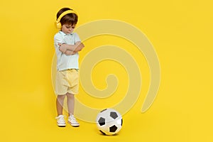 Full lengh photo of sad little boy with soccer ball wearing headphone over yellow background.