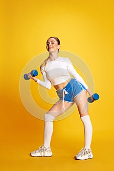 Full lenght image of young woman in training pumping up muscles of hands and legs looking at camera over yellow