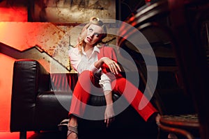 Full-lenght photo of young blonde woman in red jacket looking away sitting on leather sofa