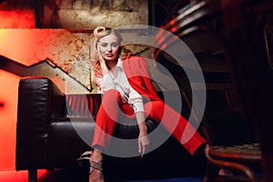 Full-lenght photo of young blonde woman in red jacket looking at camera sitting on leather sofa