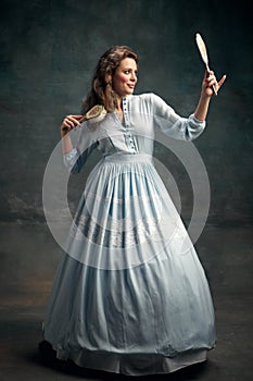 Full-lenght portrait of charming aristocratic woman wearing blue historical dress holding vintage mirror in hand and