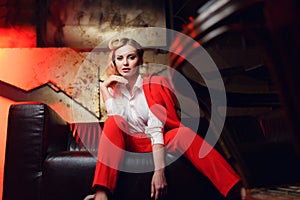 Full-lenght portrait of young blonde woman in red jacket sitting on leather sofa