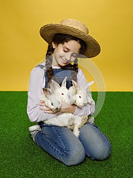 Full lenght view of a little girl in a jeans and straw hat holding a real three rabbits, isolated yellow background.