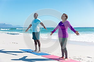 Full length of african american retired senior couple practicing yoga at beach against blue sky