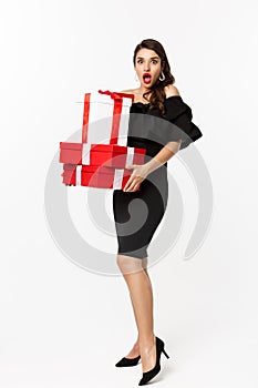 Full length of attractive female model in glamour dress, red lips, holding christmas gifts and looking amazed, receive
