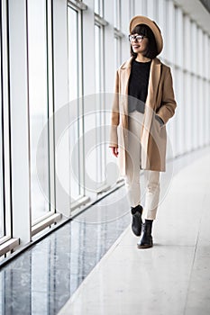 Full length of beautiful woman wearing glasses and beige coat walking in office neaw windows