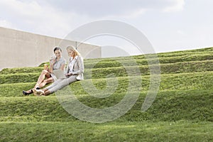 Full length of beautiful young businesswomen using laptop while sitting on grass steps against sky