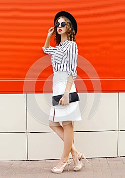 Full-length beautiful young woman model wearing white striped shirt, black handbag clutch, round hat, skirt on city street over