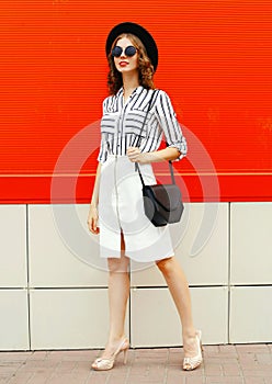 full-length beautiful young woman model wearing white striped shirt, black handbag clutch, round hat, skirt on city street over