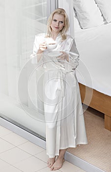 Full length of beautiful young woman in robe drinking coffee at balcony doorway