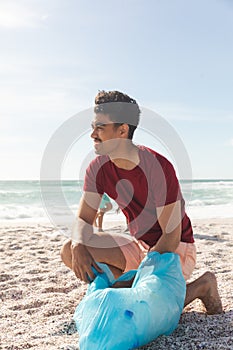 Full length of biracial man collecting garbage in bag at beach against sky with copy space