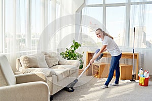 Full length body portrait of young woman in white shirt and jeans cleaning carpet under sofa with vacuum cleaner in living room.