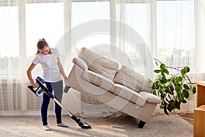 Full length body portrait of young woman in white shirt and jeans cleaning carpet with vacuum cleaner under sofa in living room.