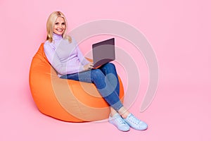 Full length body size photo of old woman using laptop smiling in orange beanbag isolated on pastel pink color background