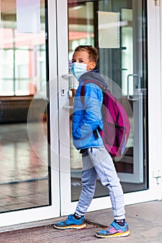 Full length boy wearing protective mask is trying to open the school door. Behind the backpack Schoolboy look at camera