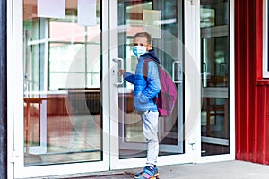 Full length boy wearing protective mask is trying to open the school door. Behind the backpack Schoolboy look at camera