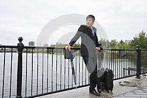 Full length of businessman with luggage leaning on railing along river