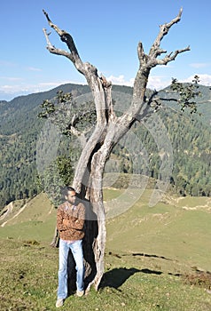 Full length of a charming south asian young guy looking sideways posing with arms crossed while leaning against tree in hilly area