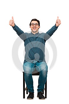 Full length cheerful and successful businessman, seated on a chair, shows thumbs up gesture, looking to camera isolated on white