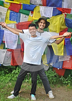 Full length of cheerful two male friends looking at camera, spreading hands against tibetan buddhist prayer flags
