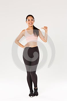 Full length of confident smiling asian female athelte in sportsbra and leggigns, fitness girl pointing at herself, lead