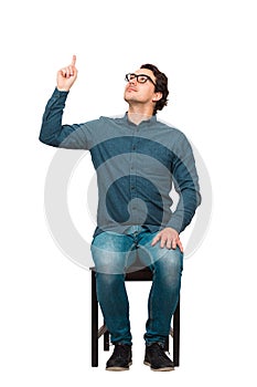 Full length contented businessman, seated on a chair, pointing forefinger looking up satisfied, isolated on white background.