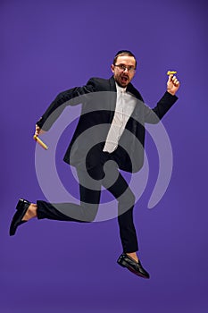 Full-length of emotional Jewish man in his 30s wearing black suit, holding noisemaker and jumping against purple studio